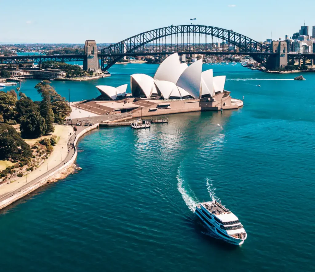 Aerial view of Sydney Harbour, featuring the iconic Opera House with its white sails, the Harbour Bridge, and a boat navigating the blue waters.