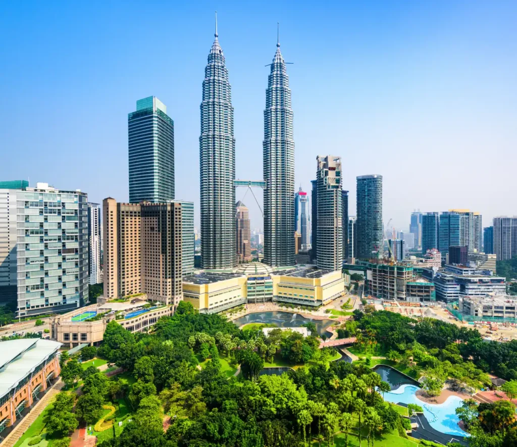 Kuala Lumpur skyline featuring the Petronas Twin Towers surrounded by modern skyscrapers. Lush green park with winding paths in the foreground. Clear, sunny day.