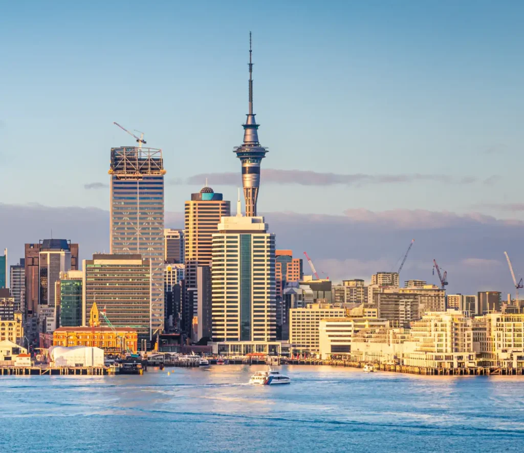 Sunlit cityscape of Auckland with the Sky Tower dominating the skyline, surrounded by modern buildings, cranes, and a boat on the harbor. Calm, urban vibe.