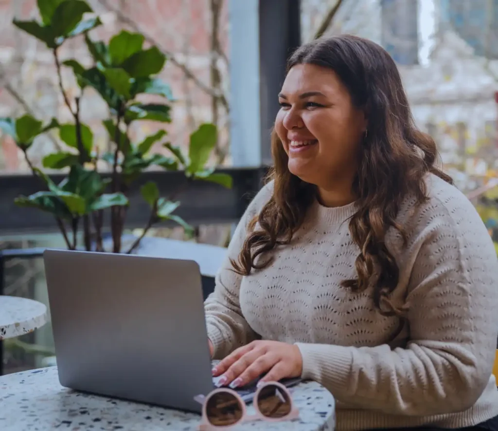 A woman with long hair smiles while using a laptop at a cafe. She's seated at a speckled table with sunglasses beside her, surrounded by green plants.