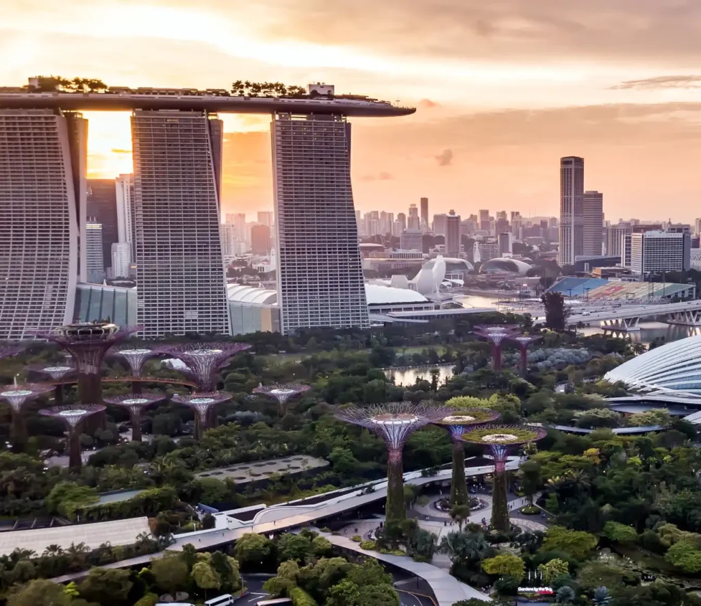 A stunning sunrise view of Singapore's Marina Bay Sands and Gardens by the Bay, featuring futuristic Supertree structures amid lush greenery. Urban skyline in the background.