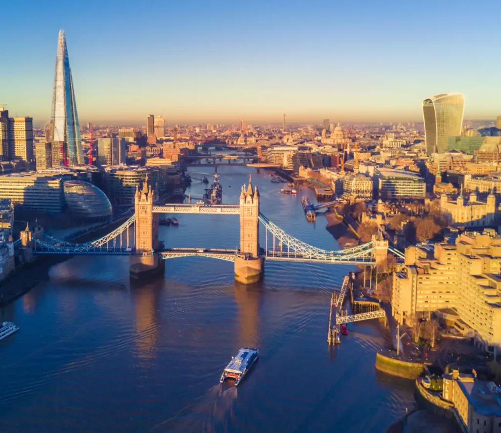 Aerial view of London's Tower Bridge over the River Thames at sunrise, with skyscrapers like The Shard and cityscape bathed in warm, golden light.