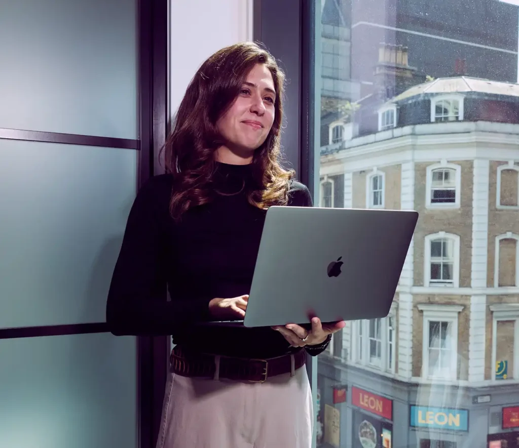 A woman in a black top holds a laptop, smiling softly while standing by a window. Outside, a city scene with historic buildings is visible.