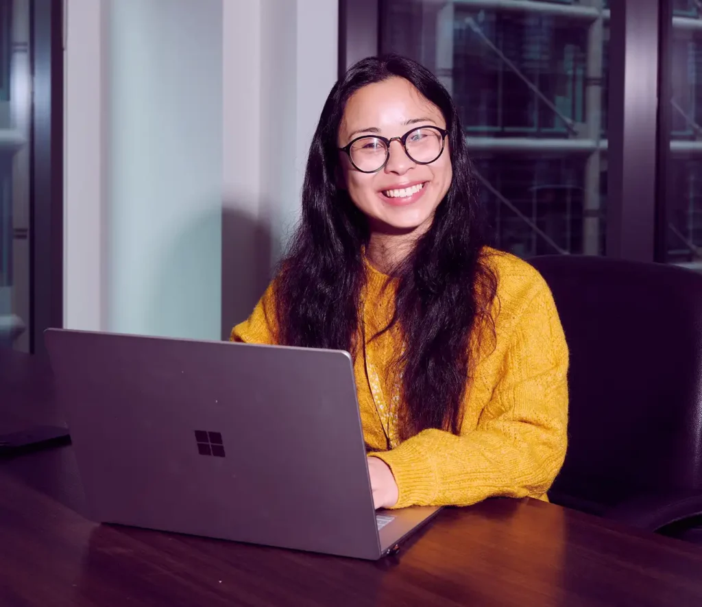 Smiling person with long dark hair and glasses, wearing a yellow sweater, working on a laptop at a desk in a modern office setting.