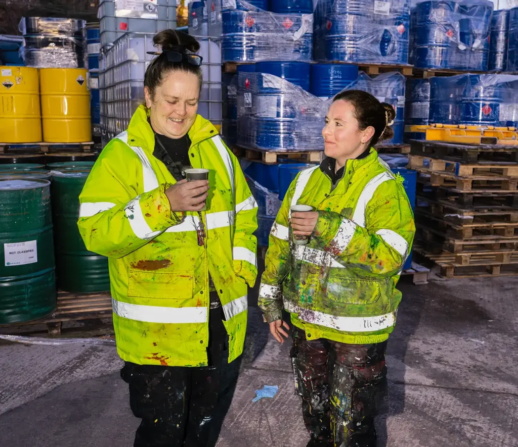 Two people in high-visibility jackets, splattered with paint, enjoy a coffee break in an industrial setting surrounded by stacked barrels. They appear relaxed and cheerful.