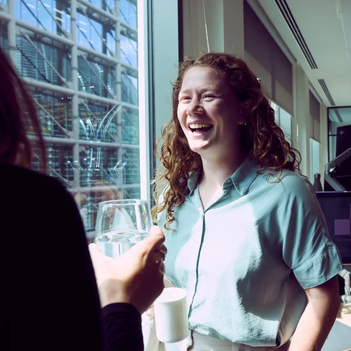 A woman with curly hair in a light blue shirt laughs while holding a cup by a sunny office window. Another person, holding a glass, faces her.