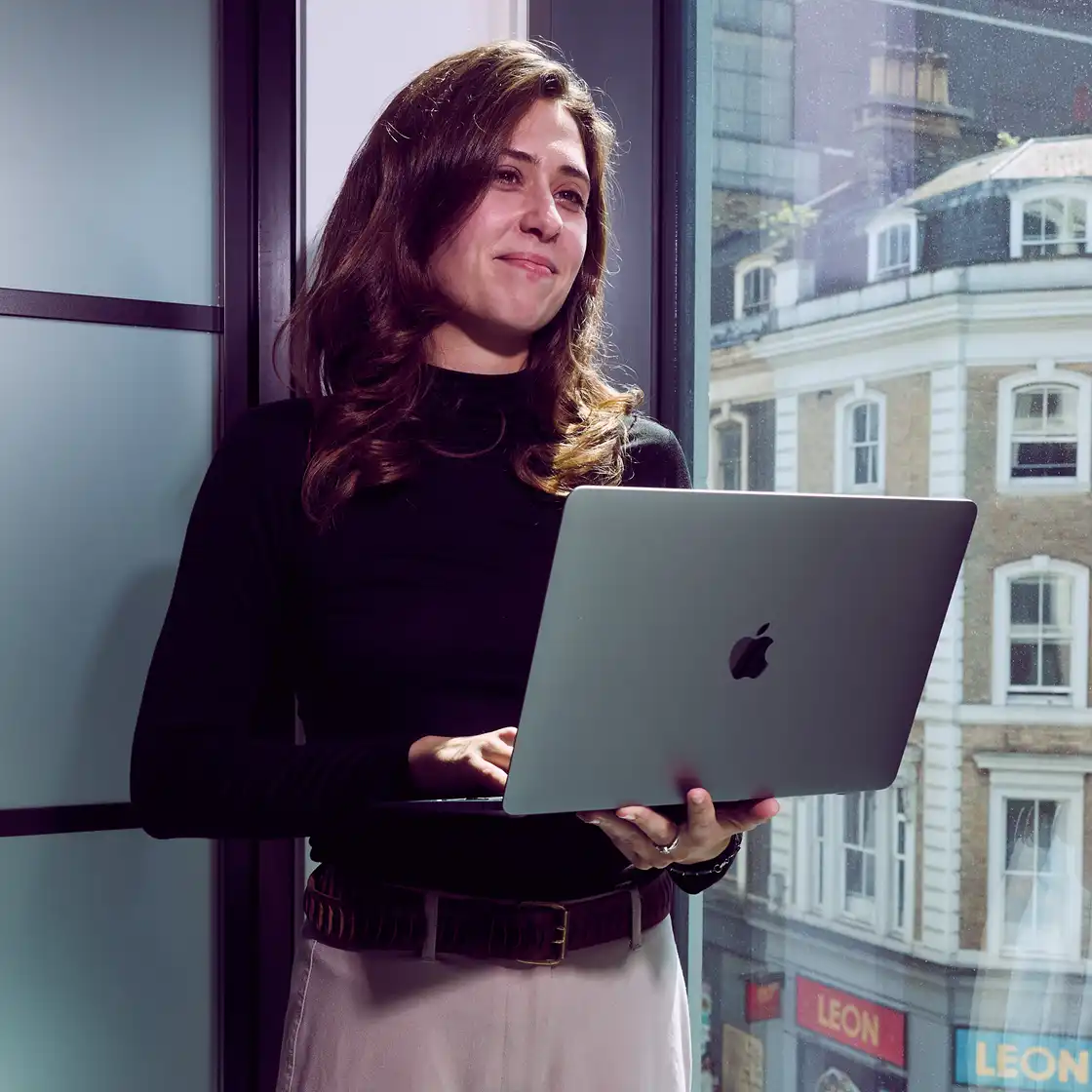A woman in a black turtleneck holds a laptop, standing by a window with a city view. She appears focused and content in a modern office setting.