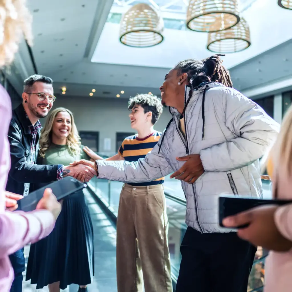 A diverse group of people joyfully greet each other indoors. Two individuals shake hands, smiling. Modern lighting and natural light create a warm atmosphere.