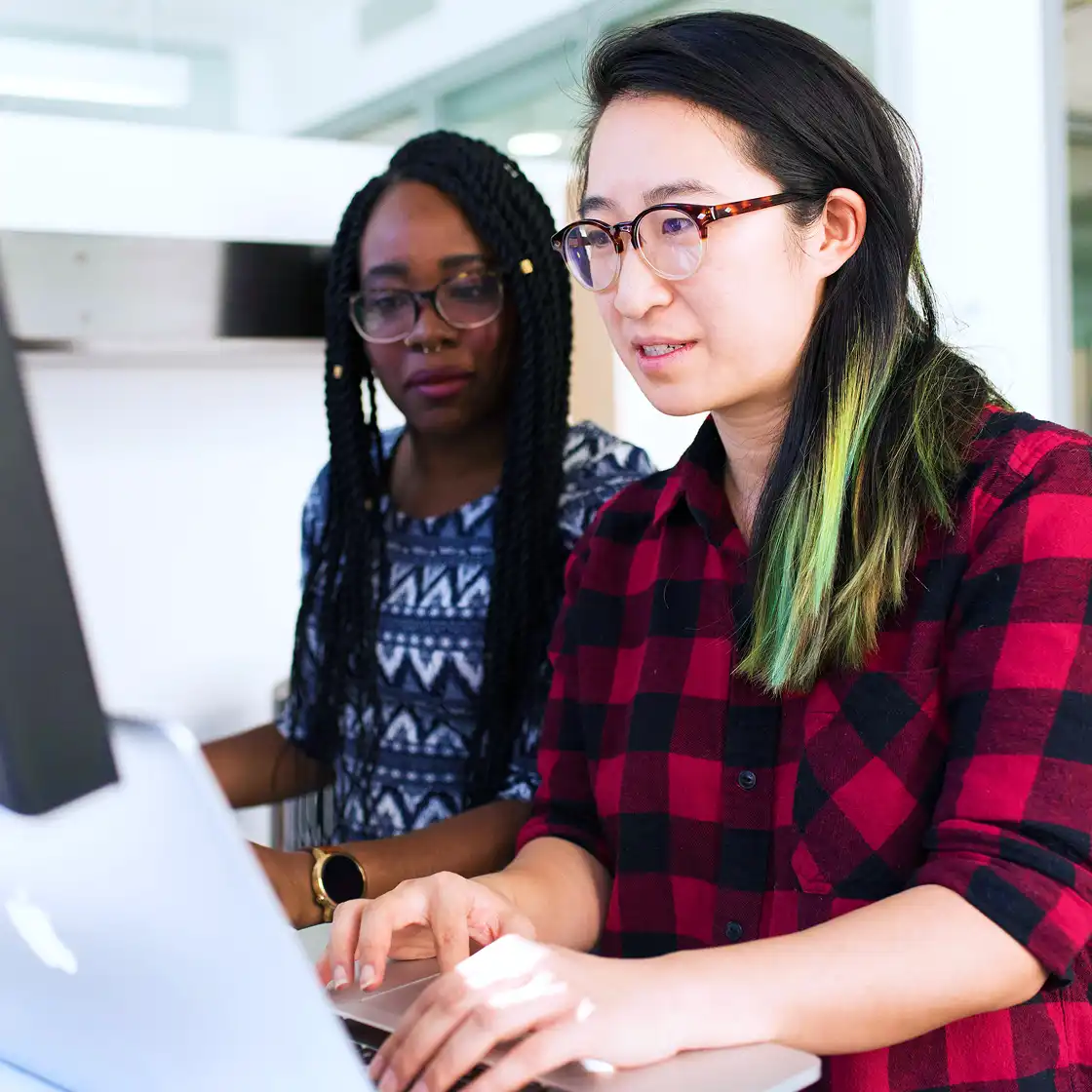 Two women collaborate on a laptop; one wears glasses and a red plaid shirt, the other wears a patterned sweater. The atmosphere is focused and professional.