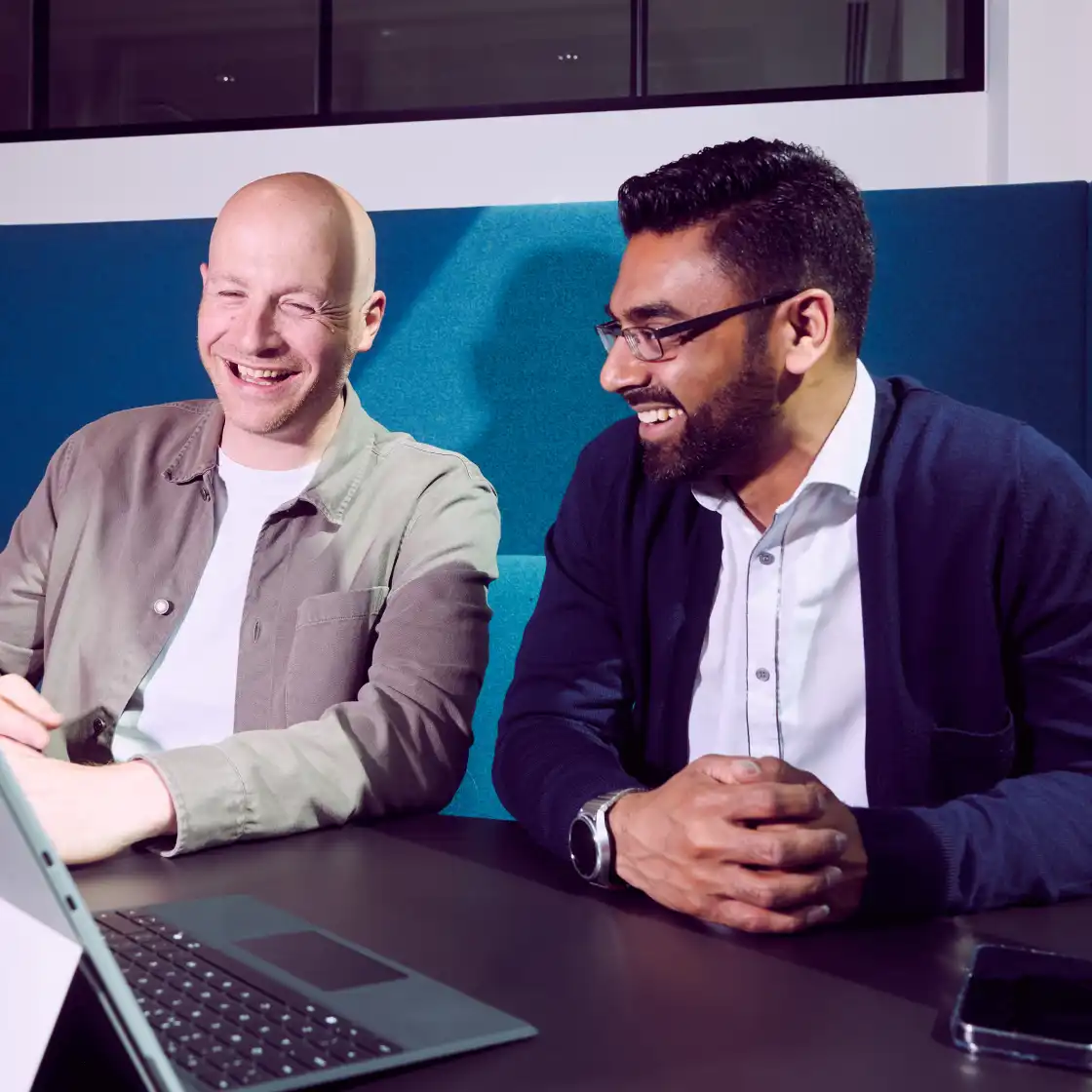 Two men sitting at a table, smiling and engaged in conversation. They are in a bright office setting with a laptop in front of them, conveying a positive, collaborative atmosphere.