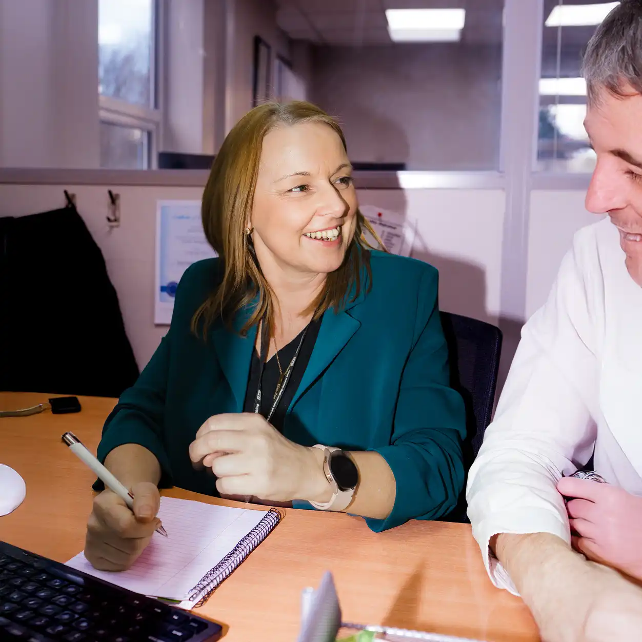 Smiling woman in a teal blazer writing in a notebook and chatting with a man in a white shirt at an office desk, conveying a friendly and collaborative atmosphere.