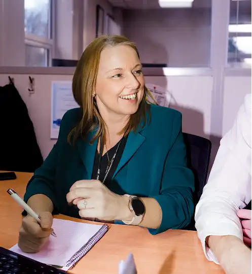 Smiling woman with light brown hair wearing a teal blazer sits at a desk, holding a pen over a notepad. She appears engaged and attentive.