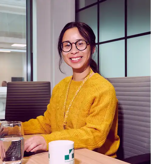 Smiling woman wearing a yellow sweater and glasses sits at a desk in a modern office, with a laptop, glass of water, and mug in front of her.