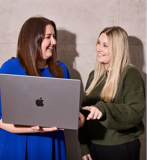 Two women smile and discuss something on a laptop. The woman on the left wears a blue dress, and the one on the right wears a green sweater.