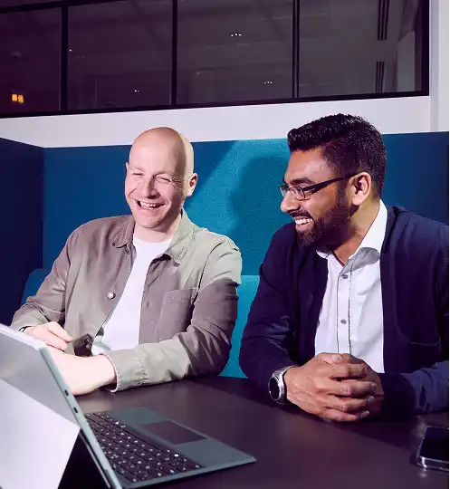 Two men in business attire sit at a table, smiling and looking at a laptop screen in a modern office. The atmosphere is collaborative and positive.