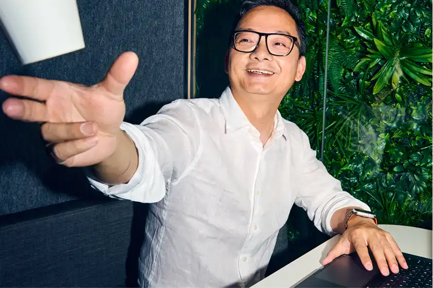 Smiling man in glasses and a white shirt reaching for a levitating cup, seated at a desk with a laptop, against a lush green plant wall background.