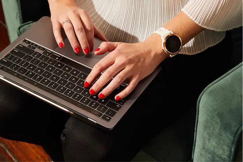 Hands with red nail polish type on a laptop keyboard. The person wears a white blouse and a smart watch, sitting on a green chair, suggesting focus.