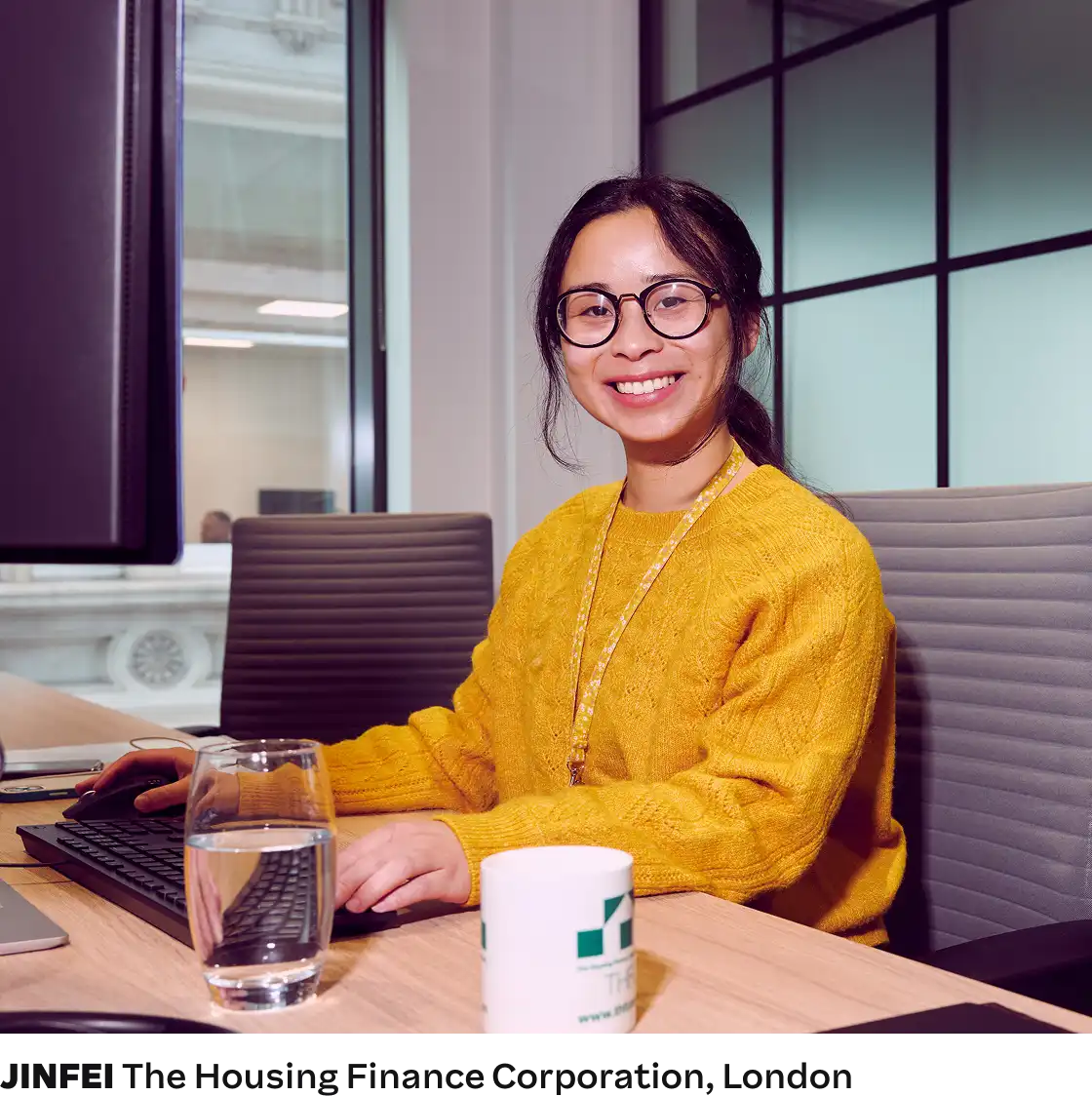 A person wearing glasses and a yellow sweater sits at a desk, smiling. A glass of water and a branded mug are in front, with a modern office setting.