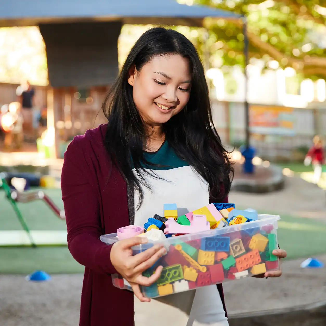 A woman smiling while holding a box of colorful toy blocks. She stands outdoors in a sunlit playground, conveying a joyful and warm atmosphere.