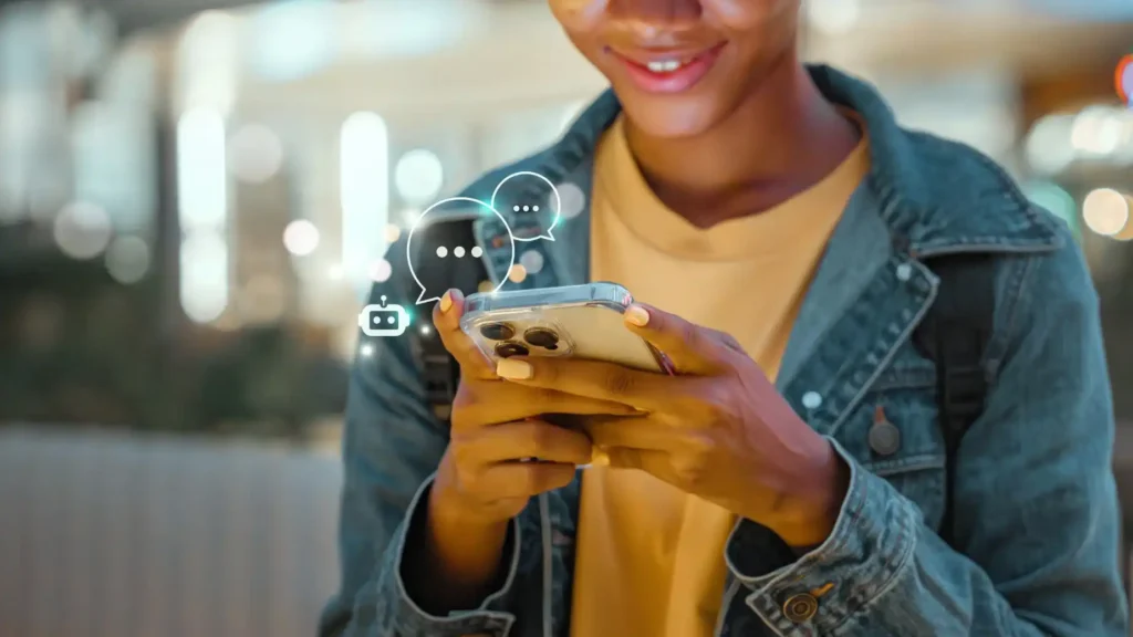 A person smiles while looking at a smartphone, with digital chat icons floating above. They wear a denim jacket, and the background is softly blurred.