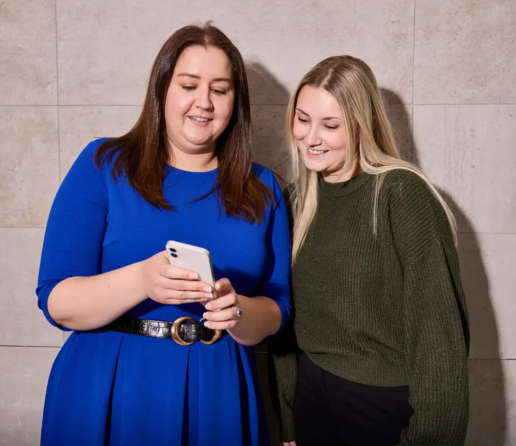 Two smiling women stand against a beige tiled wall. One in a blue dress holds a smartphone; the other in a green sweater looks at the screen, conveying a friendly mood.