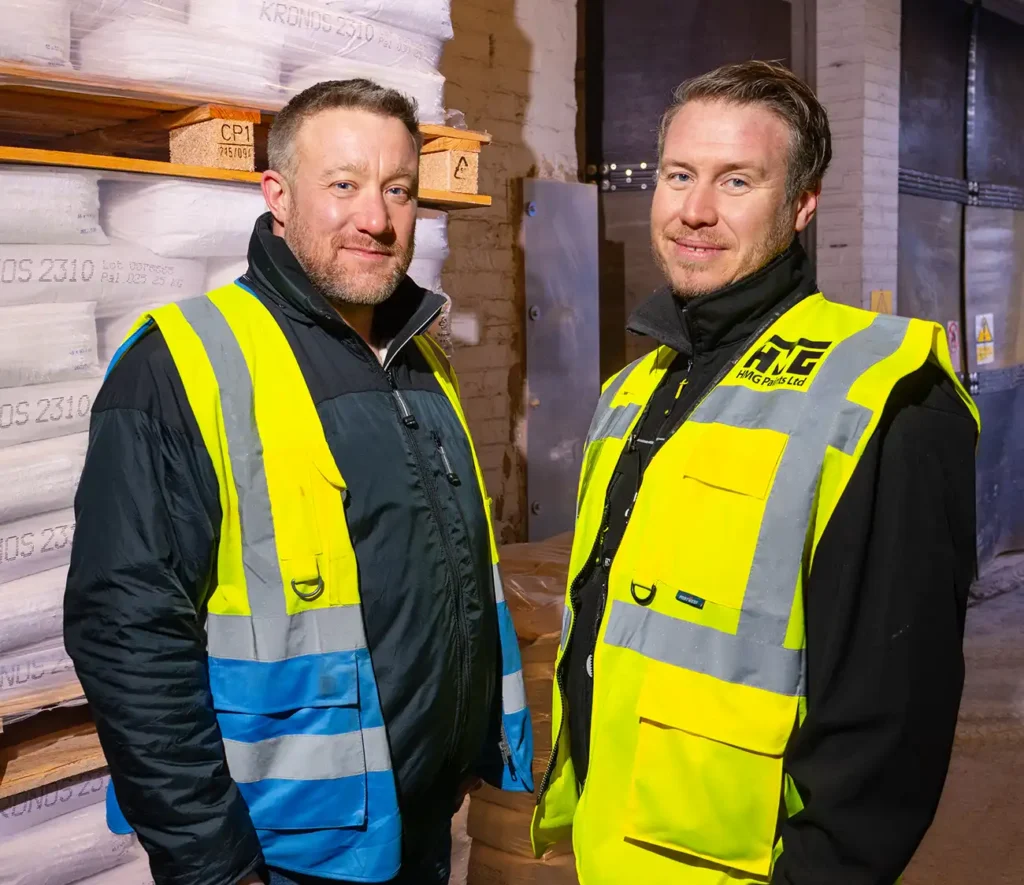 Two men in high-visibility vests stand smiling in a warehouse. Stacked bags of goods are in the background, conveying a work environment.