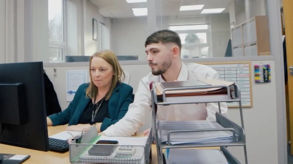 A man and a woman in an office work intently at a computer. The room is bright with organized paperwork. They convey focus and collaboration.