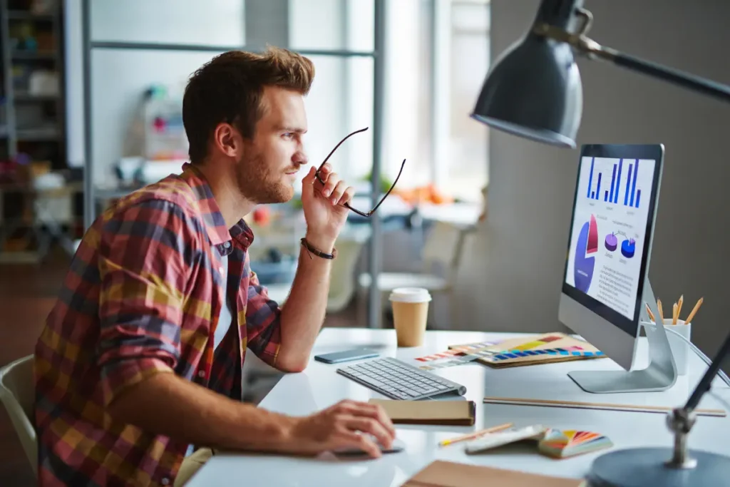 Man analyzing data charts on desktop computer while working at modern office desk.