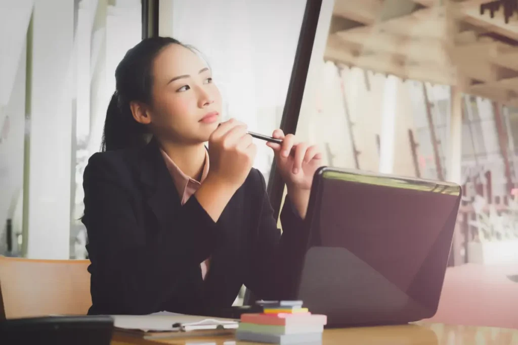 Businesswoman sitting at a desk with a laptop, holding a pen and looking thoughtfully out the window.