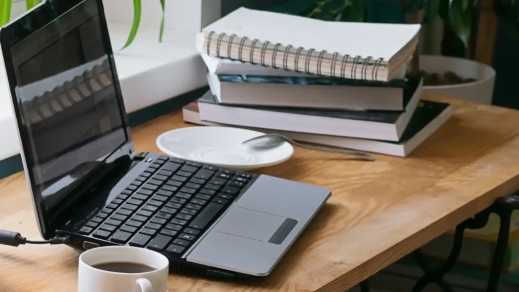 A laptop sits on a wooden desk beside an empty cup and saucer. A stack of books with a spiral notebook on top is in the background, creating a calm study atmosphere.