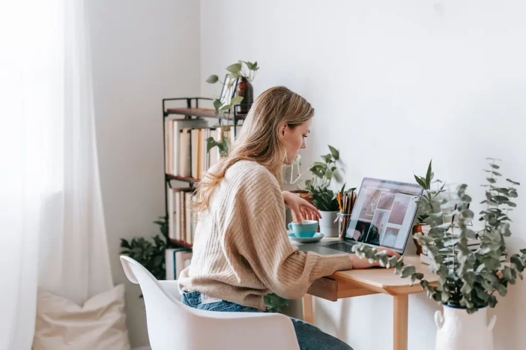 Woman working from home at desk with laptop surrounded by indoor plants and books.