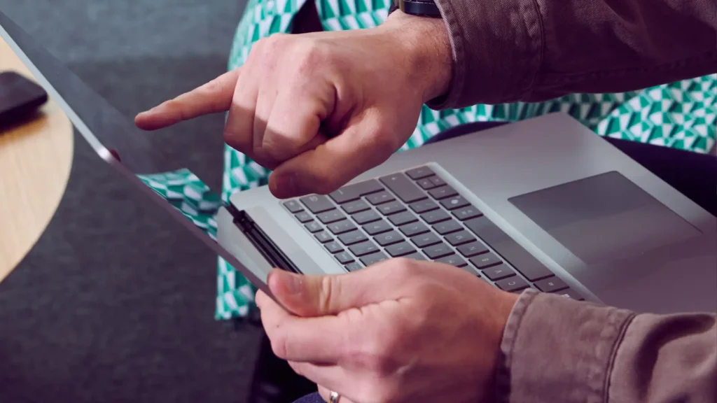 A person uses a laptop, pointing at the screen with focus. The setting is casual, indicated by a patterned cloth in the background, conveying productivity.
