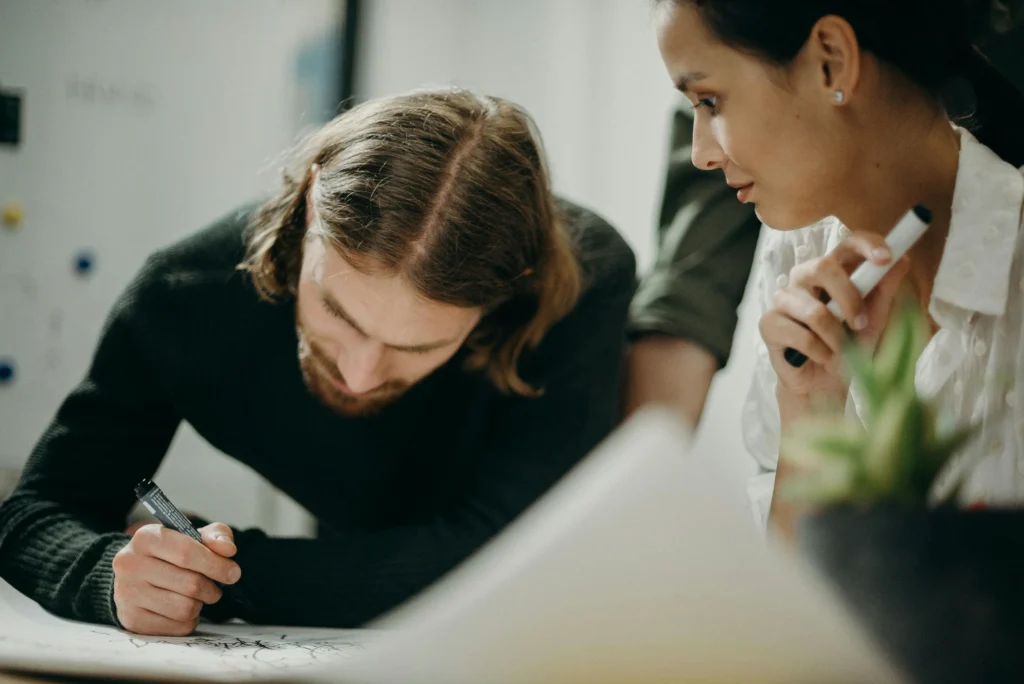 Close-up of two colleagues, a man and a woman, leaning over a desk while collaborating and making marks on a large sheet of paper.