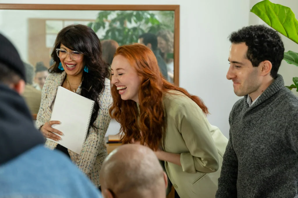 A diverse group of colleagues laughing together in a brightly lit office setting, with one woman holding a white notebook.