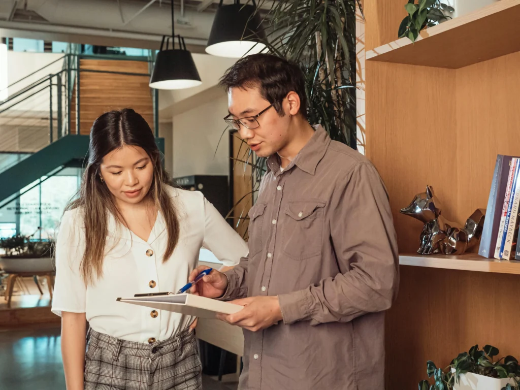 A male and female colleague standing in a modern, plant-filled office space, reviewing a document on a clipboard.