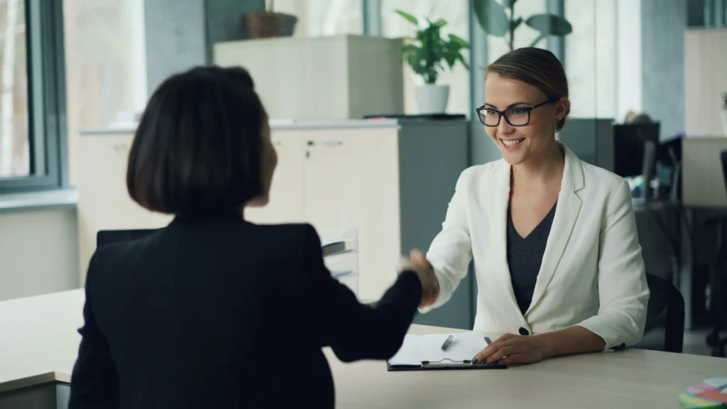A manager and a new female employee shaking hands in an office, symbolizing the start of the onboarding process and the tracking of employee onboarding metrics and KPIs.