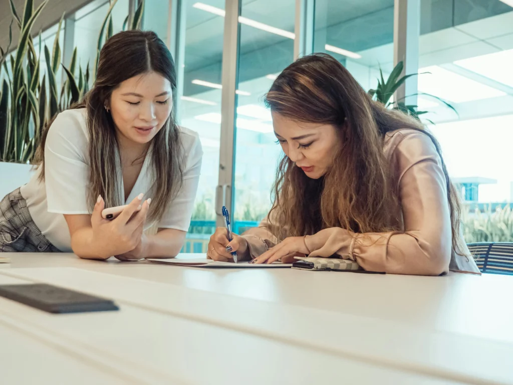 Two Asian women working together at a desk; one is writing and signing a document, while the other holds a mobile phone.