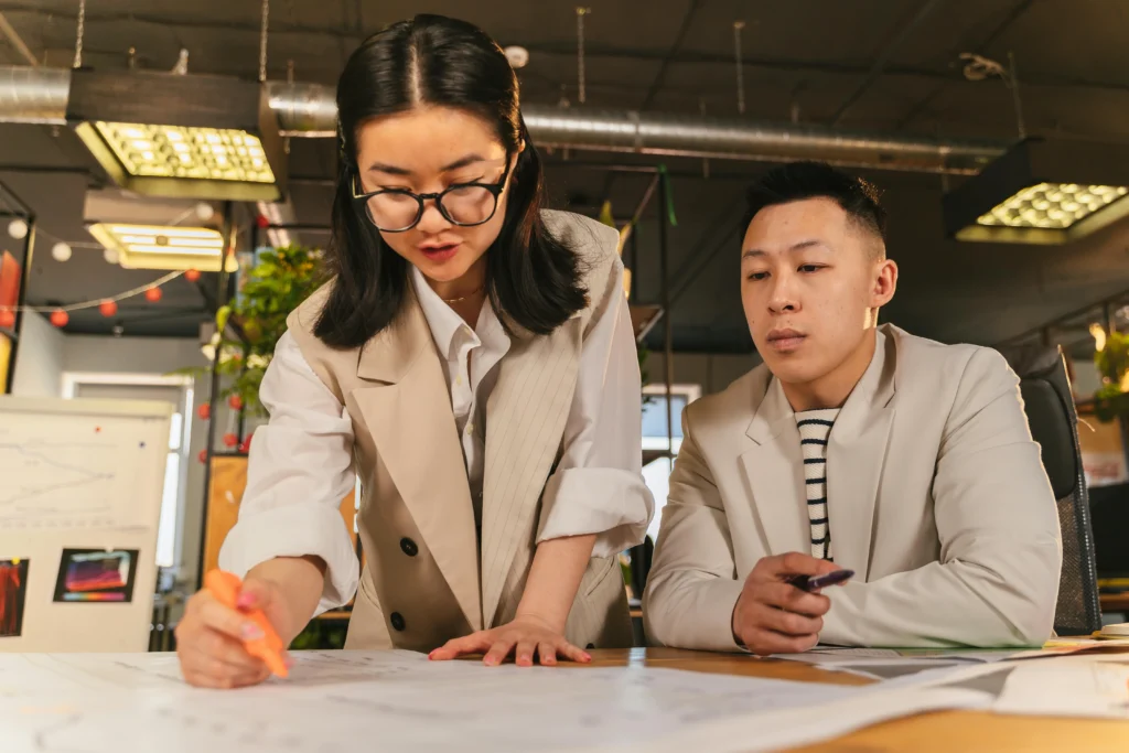 Two colleagues, a woman in glasses and a man, standing over a large blueprint or architectural plan, marking it up with pens.