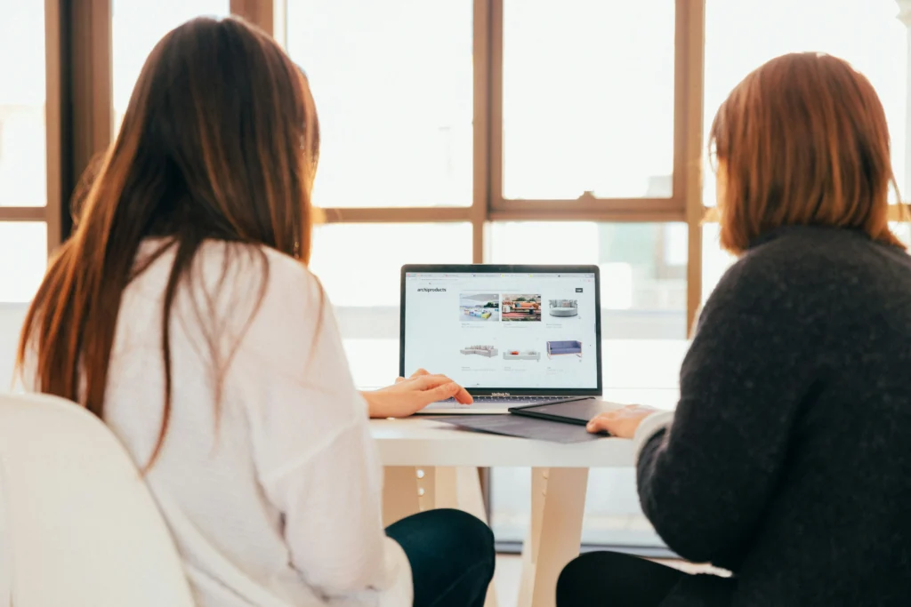 Two people sitting at a white table, reviewing product listings and design elements on a laptop displaying an e-commerce website.