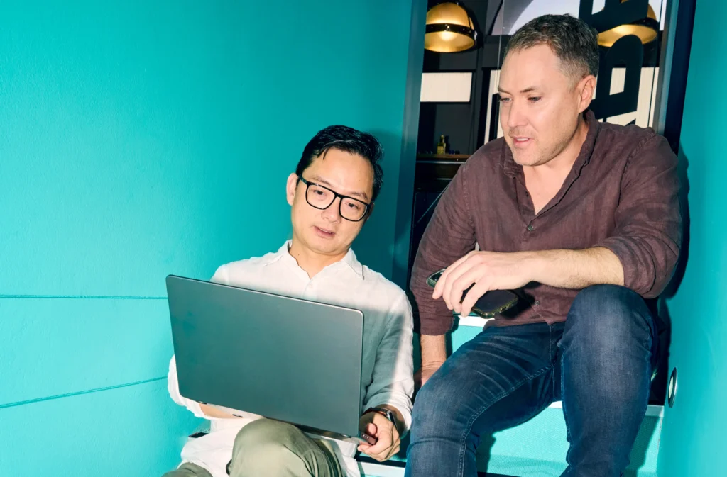 Two men sitting on bright teal stairs, looking at a laptop screen and collaborating in an office setting.