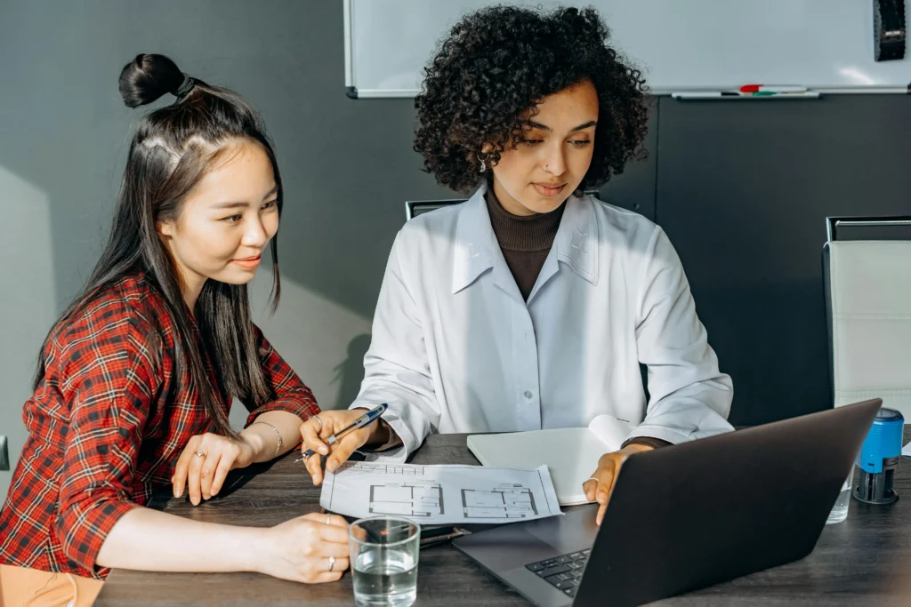 Two diverse professional women collaborating on a floor plan document while looking at a laptop in a modern office.
