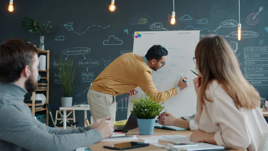 A man in a mustard sweater writes on a flip chart during a team meeting in a modern office with a chalkboard wall. Colleagues watch attentively.