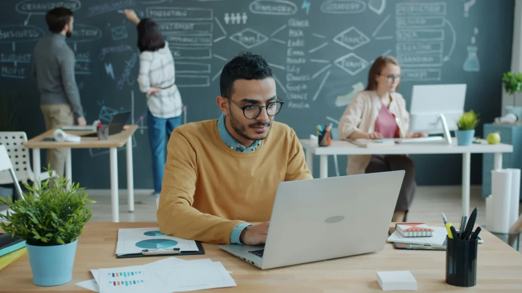 A focused man in glasses works on a laptop at a desk with plants. Behind, two people write on a chalkboard. The environment is collaborative and studious.