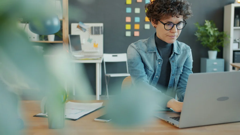 A person with curly hair and glasses works intently on a laptop in a cozy office, with greenery in the foreground and colorful sticky notes on the wall.