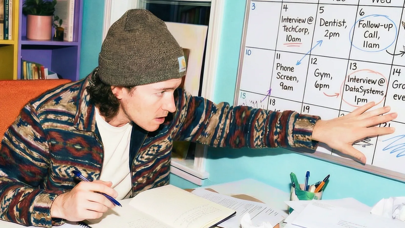 A man in a beanie is engaged in writing on a calendar, planning his upcoming events and appointments.