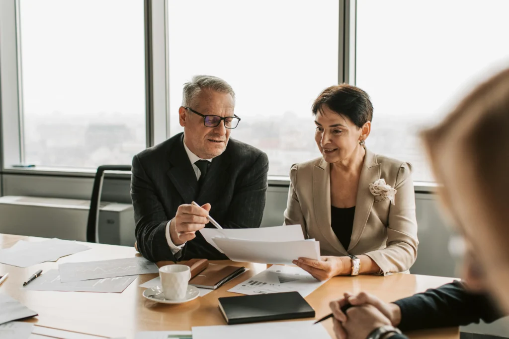 A businessman and businesswoman sitting at a desk in a bright office while reviewing financial documents together.