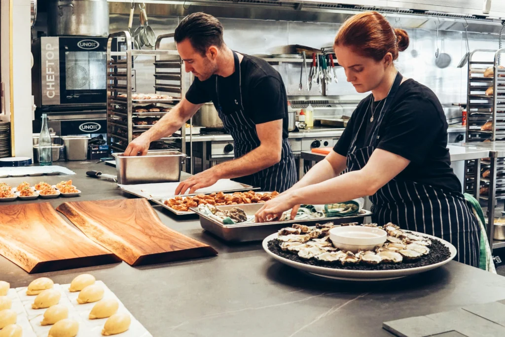 Two chefs plating food in a professional kitchen with trays and cooking equipment