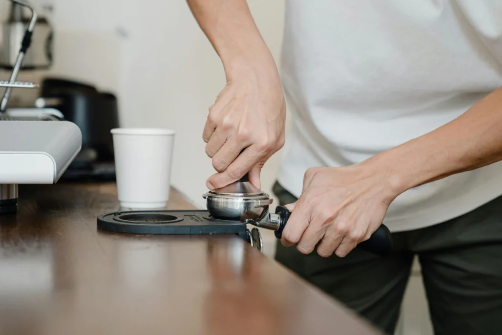 Person tamping ground coffee into portafilter on wooden counter near espresso machine