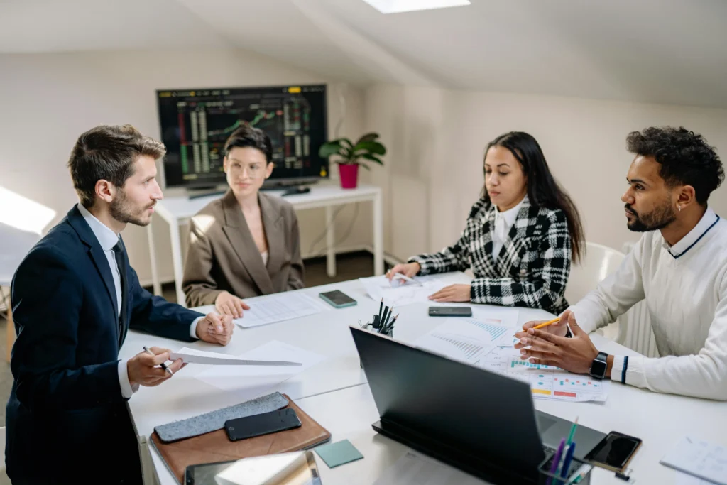 A group of four diverse professionals sitting around a white table discussing printed charts and data.