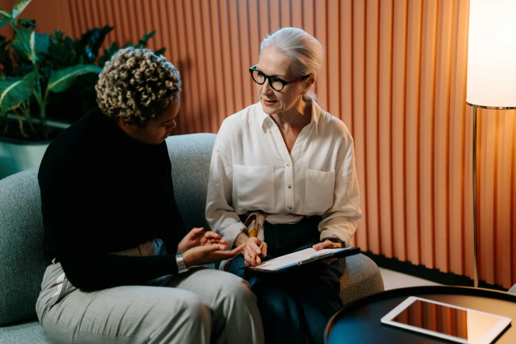 A senior woman with glasses reviewing a document on a clipboard while seated on a sofa talking to a younger colleague.